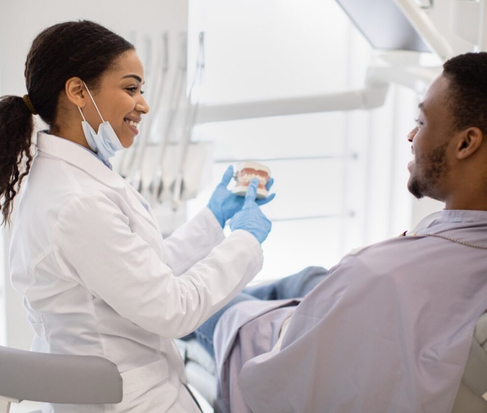 Smiling Black Stomatologist Lady Consulting Male Patient In Clinic, Holding Plastic Jaw